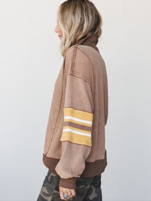 A side-profile shot of a washed mocha pullover sweatshirt with a vintage varsity stripe on the sleeve, styled with camo pants — the kind of top that looks like it's been loved for years but just arrived.
