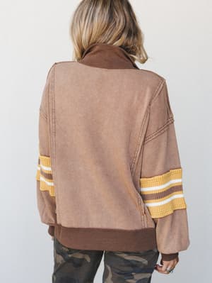 Shot from the back, a model wears a washed mocha sweatshirt with varsity-stripe sleeve detailing and camo pants — the kind of lived-in, effortlessly cool combo that feels like it came together on a Sunday without trying.