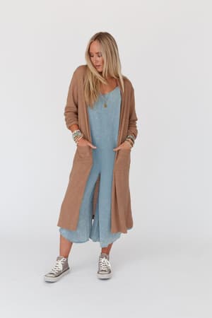 A woman with long blonde hair wears a light blue dress, gray Converse sneakers, and The Nest Jayde Waffle Knit Cardigan in taupe. She stands against a plain white background, looking slightly downward with her hands in her cardigan pockets.