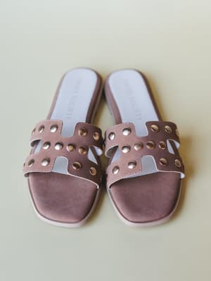 A pair of dusty mauve slide sandals with cutout H-strap detailing and gold stud embellishments, shot overhead on a soft sage-cream surface — the kind of sandal that quietly pulls an outfit together.