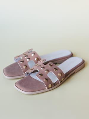 A pair of dusty mauve slide sandals with a cutout H-strap and gold stud embellishment, shot on a soft cream surface — the kind of shoe that goes with everything but still has something to say.