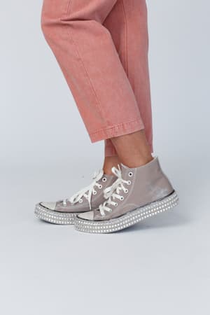 A person wears mocha Chantel Studded High Top Sneakers with thick white soles and rhinestone details, paired with light pink pants, standing against a plain light background.