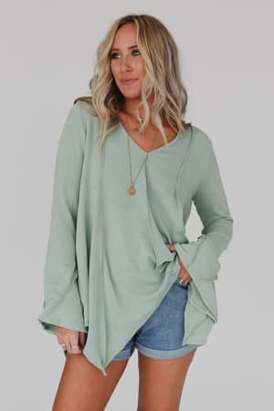 A woman with long blonde hair wears the Golden Hour Long Bell Sleeve Top in sage, paired with denim shorts and a gold necklace, standing against a plain light gray background with one hand in her pocket.