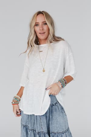 A woman with long blonde hair wears The Nest Your Favorite Slub Tee Shirt in Natural, a tiered denim skirt, colorful beaded bracelets, and layered necklaces. She stands with one hand in her skirt pocket against a light background.