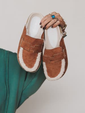 Green / Brown suede loafers with white fur lining held by a person against a neutral background