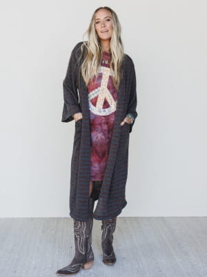 Woman wearing a long dark coat over a tie-dye shirt with a peace symbol, standing against a white wall.