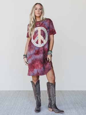 Woman wearing a tie-dye dress with a peace symbol and cowboy boots on a plain background