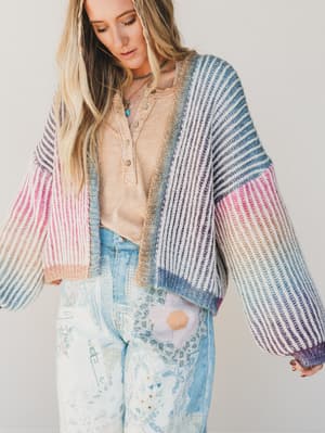Woman wearing a colorful striped cardigan over a beige shirt and floral pants on a plain background