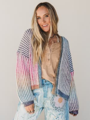Woman wearing a colorful striped cardigan over a beige shirt and floral pants on a plain background