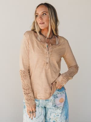 Woman wearing a beige long-sleeve top with lace cuffs and floral pants against a plain background