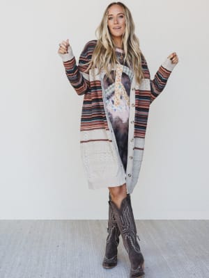 Woman wearing a long, patterned cardigan over a dress with cowboy boots on a plain background