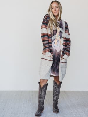 Woman wearing a long, patterned cardigan over a dress with cowboy boots on a plain background