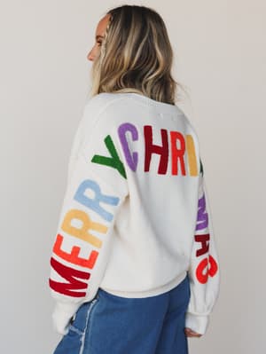 Person wearing a white sweater with colorful letters spelling 'MERRY CHRISTMAS' on a plain background
