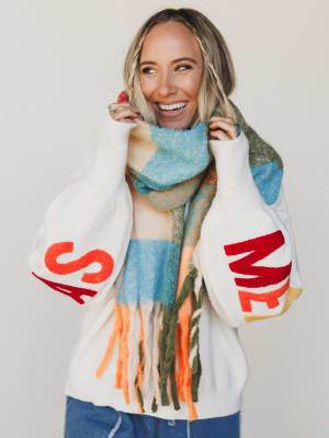 Woman wearing a colorful scarf and a white sweater with red text, smiling against a plain background