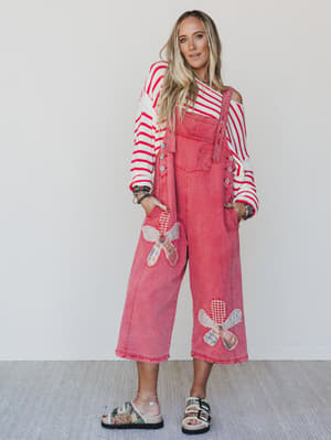 Woman wearing a pink denim overalls with floral patterns and a striped long-sleeve shirt against a white background.