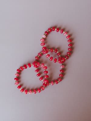 Two beaded stretch bracelets laid overlapping on a soft grey surface — red and pink stone beads separated by small gold tube beads, casual and collectible in the best way.