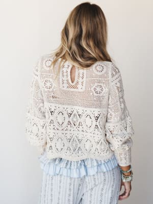 Shot from behind, a cream crochet top with intricate open-knit paneling and a keyhole back is layered over a ruffled floral midi skirt — the kind of outfit that looks like it was assembled effortlessly but definitely wasn't.
