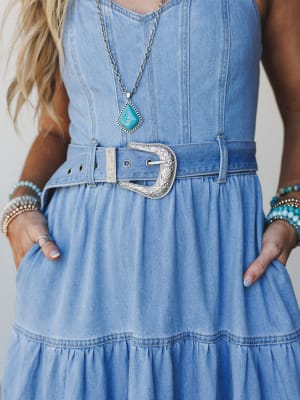 A close-up detail shot of a light wash denim tiered dress styled with a statement silver western belt buckle, turquoise stone pendant necklace, and stacked beaded bracelets — effortlessly boho with a western edge.