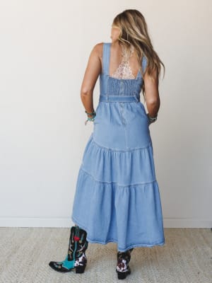 Shot from behind, a model in a tiered denim maxi dress layered over a lace bralette pairs the look with mismatched embroidered cowboy boots and stacked turquoise bracelets — effortlessly Western-boho and full of personality.