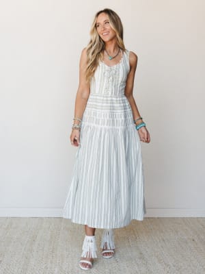 A breezy striped maxi dress with lace-up bodice detailing floats effortlessly on a smiling model styled with white fringe heels and turquoise accessories — the kind of outfit that makes a Tuesday feel like a reason to be somewhere.
