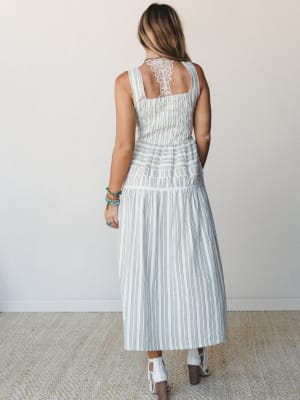 A striped midi dress shot from behind, showing off its standout lace back panel, smocked bodice, and tiered skirt — styled with white booties and a stack of turquoise bracelets for that effortless boho-meets-summer energy.