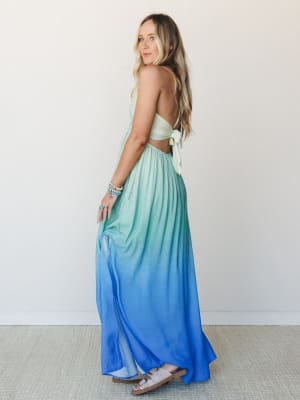 An ombre halter maxi dress flows from soft mint at the bodice into a saturated cobalt blue at the hem — shot from the back to show off the open-back tie detail, with stacked turquoise bracelets and slide sandals rounding out the look.