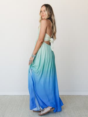 An ombre halter maxi dress flows from soft mint at the bodice into a saturated cobalt blue at the hem — shot from the back to show off the open-back tie detail, with stacked turquoise bracelets and slide sandals rounding out the look.
