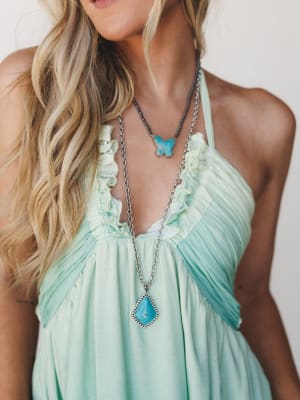 A close-up torso shot of a mint-washed halter dress with soft ruffle trim at the neckline, styled with layered turquoise stone necklaces — the kind of outfit that feels like a warm afternoon somewhere coastal.