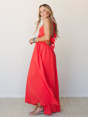 A coral-red crinkle maxi dress shot from a side-back angle, showing off the open back with tie closures and tassel detail — the kind of dress that makes you want to book a trip somewhere warm.