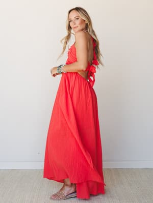 A coral-red crinkle maxi dress shot from a side-back angle, showing off the open back with tie closures and tassel detail — the kind of dress that makes you want to book a trip somewhere warm.