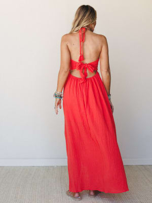 A bold cherry tomato red halter maxi dress shot from the back, showing off a dramatic open back with tie details and tassel trim — styled with stacked boho bracelets that feel totally intentional.