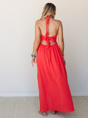 A bold cherry tomato red halter maxi dress shot from the back, showing off a dramatic open back with tie details and tassel trim — styled with stacked boho bracelets that feel totally intentional.