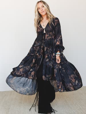 A flowy, dark floral duster coat sweeps dramatically open over black wide-leg pants with fringe boots — the whole look has that moody, free-spirited energy of someone who dresses like a feeling.