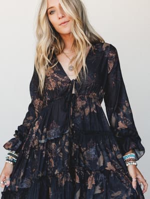 A dark floral chiffon tiered dress with billowy long sleeves and a deep v-neckline styled with layered gold necklaces and stacked beaded bracelets — moody, romantic, and the kind of outfit that makes a regular Tuesday feel like something.