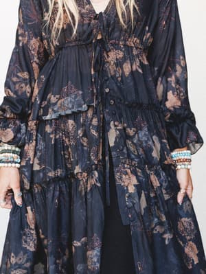 A moody, botanical-print midi dress or top-and-skirt set in deep navy and warm taupe, shot close on a model stacked in beaded bracelets — the kind of piece that feels like it was made for wandering somewhere beautiful.