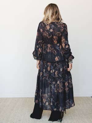 Shot from behind, a model in a dramatic dark floral maxi dress with billowy long sleeves and a tiered skirt wears it over wide-leg black pants with fringe boots — the whole look has a moody, free-spirited energy that feels equal parts concert and can