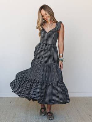 A smiling model mid-twirl in a black gingham tiered maxi dress with flutter sleeves and a sweetheart neckline, styled with layered silver necklaces, stacked bracelets, and patterned mules against a clean white wall.