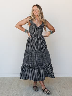 A black gingham tiered maxi dress styled with layered bracelets, a delicate silver necklace, and woven mary jane flats — the kind of outfit that looks equally at home at a farmers market or a backyard dinner party.