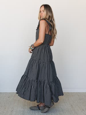 A tiered gingham maxi dress shot from behind in soft studio light — the open back, ruffle hem, and tie shoulders give it that sweet-but-not-precious energy that works just as hard at a garden party as it does at a late summer dinner.