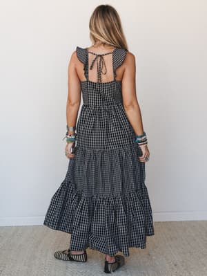 A tiered gingham maxi dress shot from the back, showing off ruffle sleeves, an open tie-back, and a full sweeping skirt — styled with stacked boho bracelets and woven flats for a look that's equal parts sweet and free-spirited.