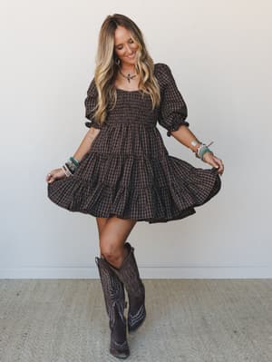 A smiling model twirls a chocolate plaid mini dress with a smocked bodice and tiered skirt, styled with tall cowboy boots and stacked beaded bracelets against a clean white wall — the kind of outfit that makes you want to spin in a parking lot.