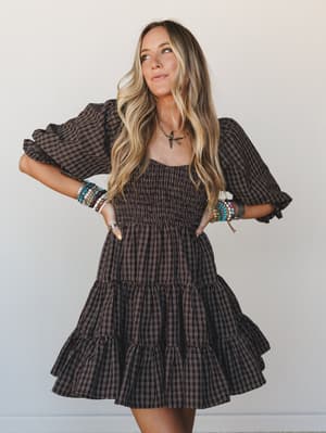 A dark chocolate plaid mini dress with dramatic puff sleeves and a tiered ruffle skirt — styled with stacked beaded bracelets and a delicate pendant necklace for that effortless boho-meets-fall energy.