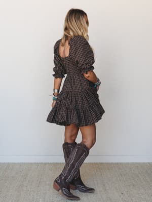 A tiered mini dress in dark chocolate plaid photographed from the back, styled with tall embroidered cowboy boots and layered boho bracelets — the kind of outfit that works just as hard at a fall market as it does at a honky-tonk.