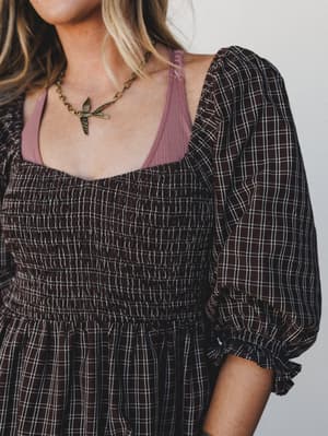 A close-up of a smocked plaid dress in deep chocolate layered over a dusty mauve cami, styled with a vintage bird pendant necklace — the kind of outfit that feels like fall in the best possible way.