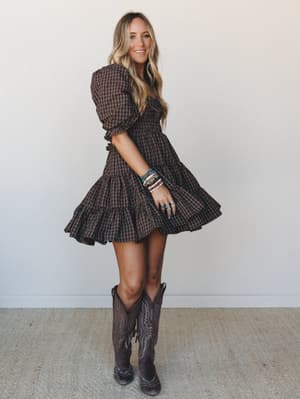 A smocked mini dress in dark chocolate plaid gets a full boho-country moment — tiered ruffles, puff sleeves, stacked beaded bracelets, and tall cowboy boots that make the whole look feel like a fall weekend with nowhere boring to be.