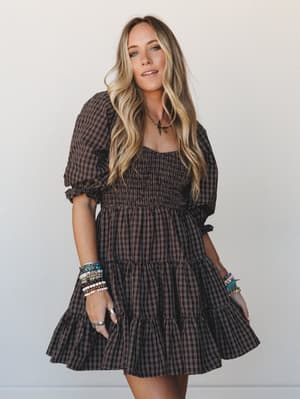 A smocked mini dress in deep chocolate plaid gets the full boho-stacked-bracelet treatment — tiered skirt, puff sleeves, and wavy hair doing all the right things against a clean studio backdrop.