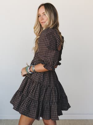 A model shot in three-quarter profile wearing a deep chocolate plaid mini dress with tiered ruffles and smocked waist, styled with a stack of colorful beaded bracelets against a clean white studio backdrop — the kind of dress that feels equally at ho
