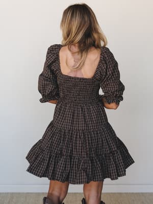 A back-facing shot of a dark chocolate plaid mini dress with dramatic puff sleeves, smocked bodice, and a flirty tiered skirt — layered over a blush lace bralette and paired with brown boots for an effortlessly fall-ready feel.