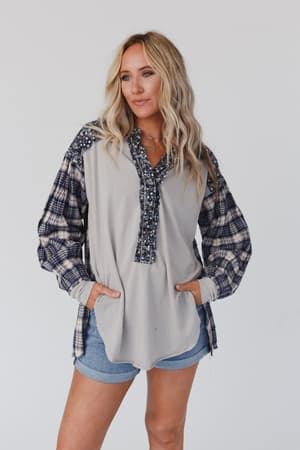 A woman with long blonde hair models the Better Together Mixed Print Top - Blue Gray, a loose boho blouse with plaid and floral patterns, paired with blue denim shorts. She stands with her hands in her shirt pockets against a white background.