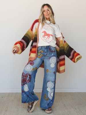 A full-body model shot layered with so much personality it practically tells a story — wide-leg patchwork jeans covered in crochet appliqués, floral embroidery, and hand-painted details paired with a white horse graphic tee and an oversized striped c